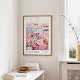 a framed poster of a colorful floral scene hanging on a wall, with a white table and a vase of flowers in the foreground.