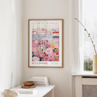 a framed poster of a colorful floral scene hanging on a wall, with a white table and a vase of flowers in the foreground.