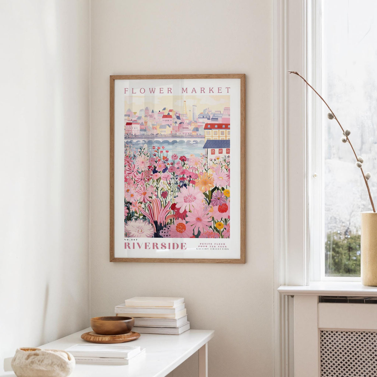 a framed poster of a colorful floral scene hanging on a wall, with a white table and a vase of flowers in the foreground.
