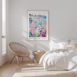 a bedroom with a bed, a woven chair, and a framed poster of a colorful floral scene hanging on the wall.