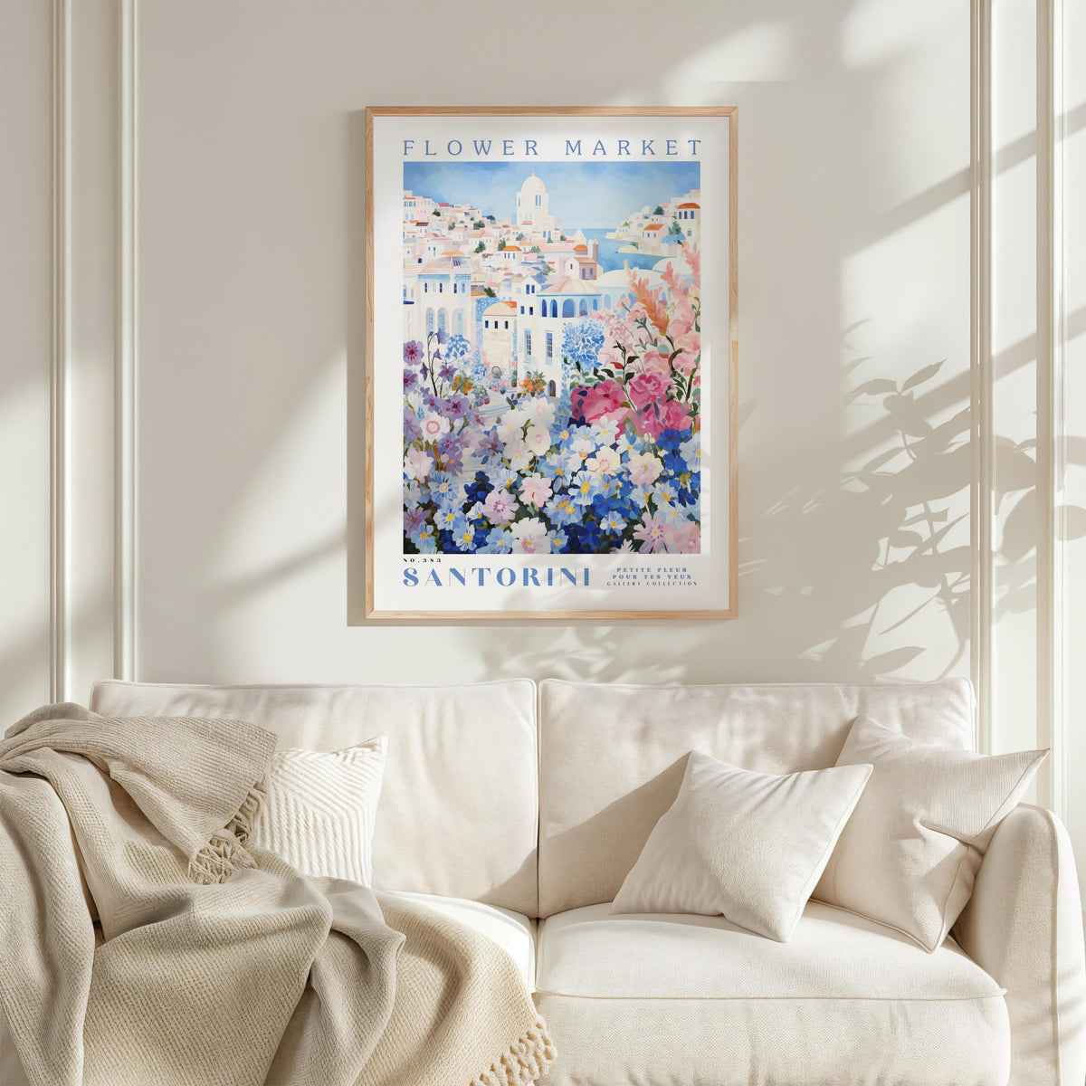 a framed poster of a flower market in Santorini, with a white sofa and a beige blanket in the foreground.