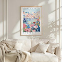 a framed poster of a colorful floral scene hanging on a wall above a white couch with a blanket draped over it.