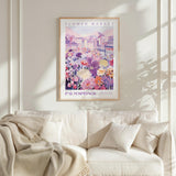 a framed poster of a colorful cityscape with flowers, hanging on a wall above a white couch with pillows.