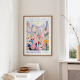 a framed poster of a colorful floral scene hanging on a wall, with a window and a small table with books and a vase in the foreground.