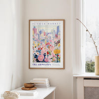 a framed poster of a colorful floral scene hanging on a wall, with a window and a small table with books and a vase in the foreground.