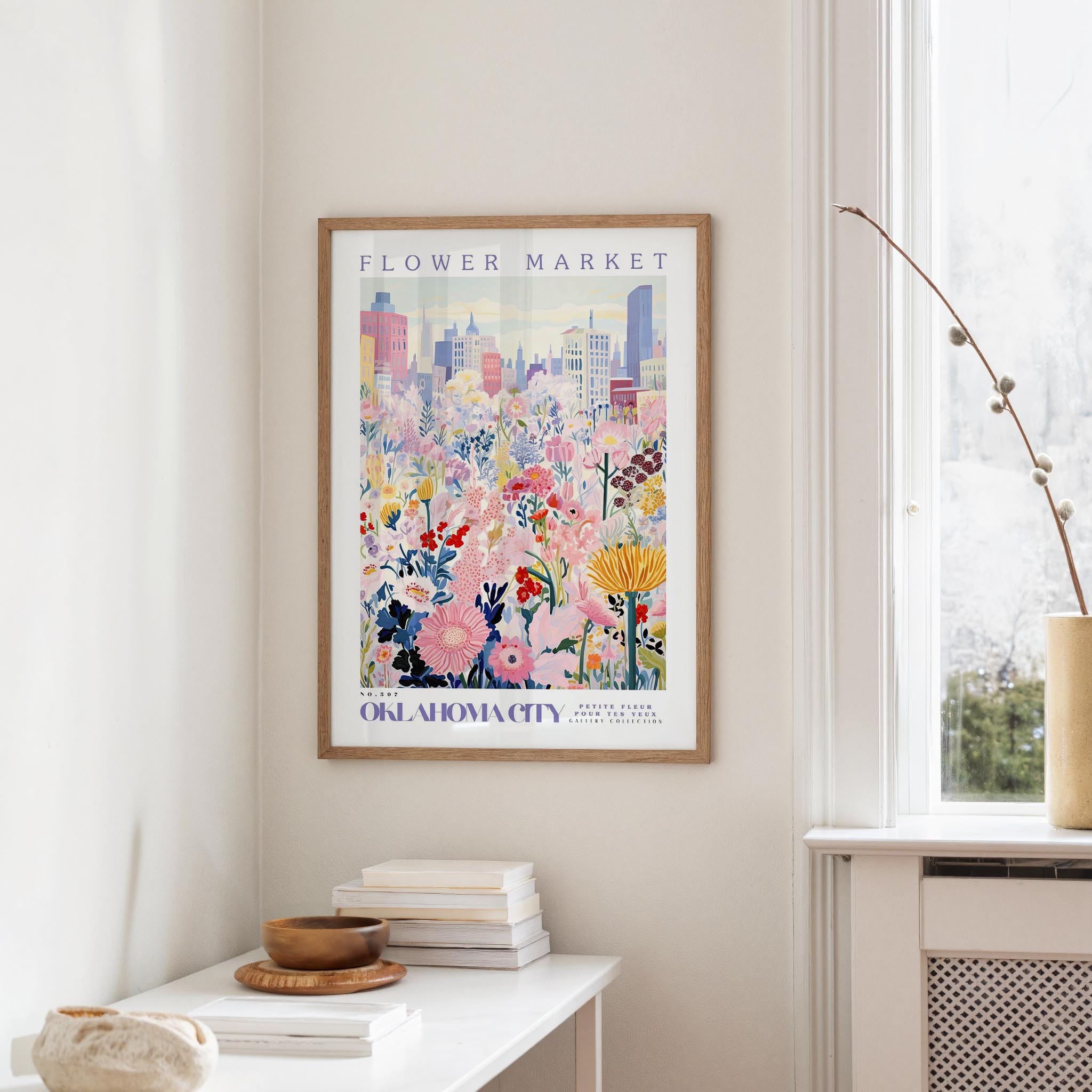 a framed poster of a colorful floral scene hanging on a wall, with a window and a small table with books and a vase in the foreground.