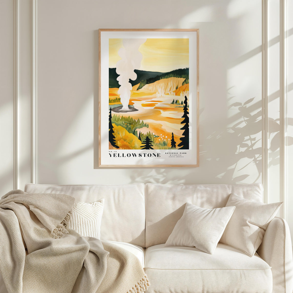a framed poster of a scenic landscape with a yellow and orange background, depicting a mountain range and a river. The poster is hanging on a wall above a white couch, which has a blanket draped over it.