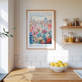 a kitchen scene with a framed poster of the "Flower Market" in Seville hanging on the wall above a wooden counter. On the counter, there is a bowl of lemons and a plant. The kitchen has white tile backsplash and shelves with various jars and containers.