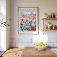 a kitchen scene with a framed poster of the "Flower Market" in Seville hanging on the wall above a wooden counter. On the counter, there is a bowl of lemons and a plant. The kitchen has white tile backsplash and shelves with various jars and containers.
