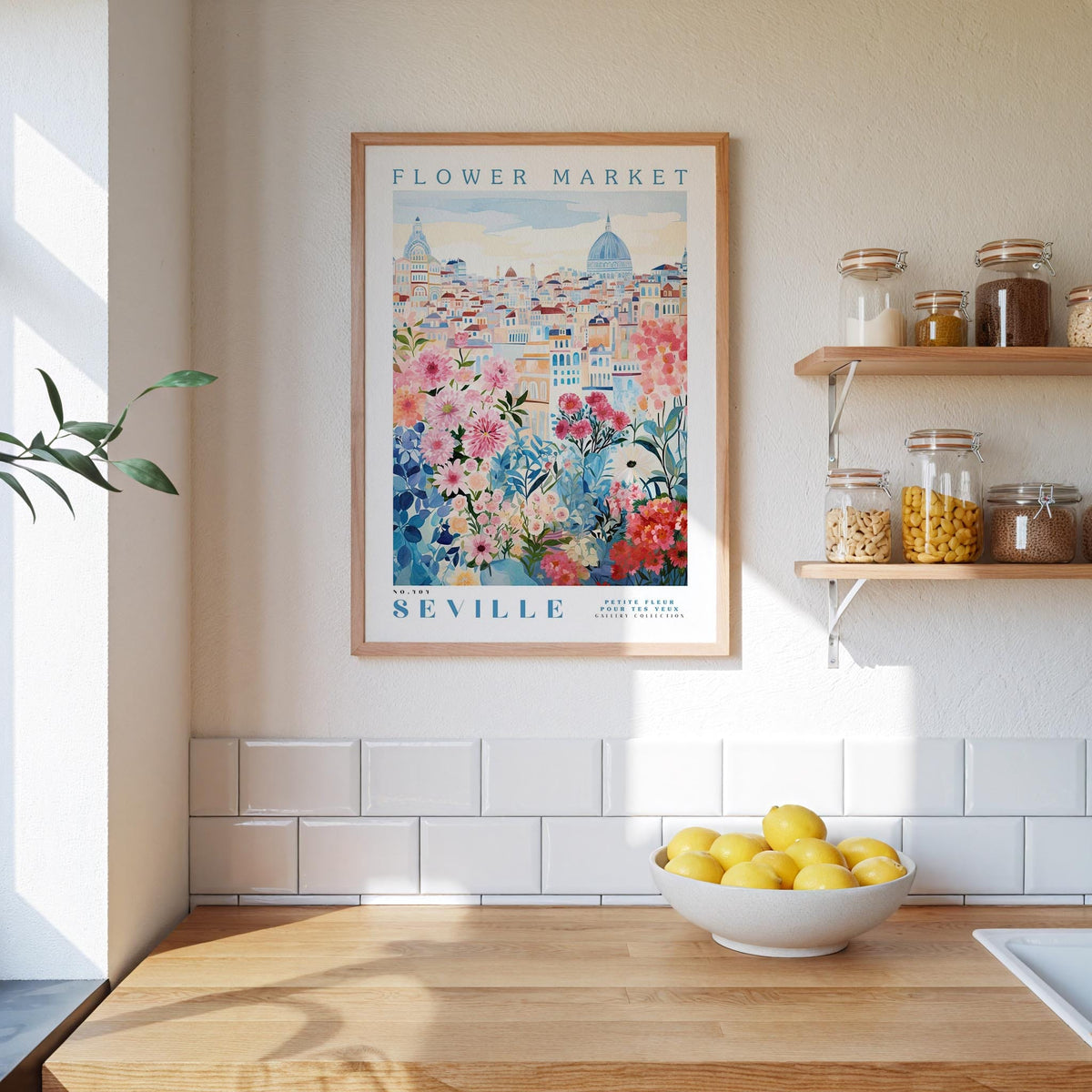 a kitchen scene with a framed poster of the "Flower Market" in Seville hanging on the wall above a wooden counter. On the counter, there is a bowl of lemons and a plant. The kitchen has white tile backsplash and shelves with various jars and containers.