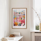 a framed poster of a colorful garden scene hanging on a wall, with a white table and a vase of flowers in the foreground.