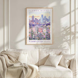 a framed poster of a cityscape with a field of flowers in the foreground, hanging on a wall above a white couch.