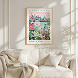 a framed poster of a cityscape with a flower market in Portland, Oregon, hanging on a wall above a white couch with a blanket draped over it.