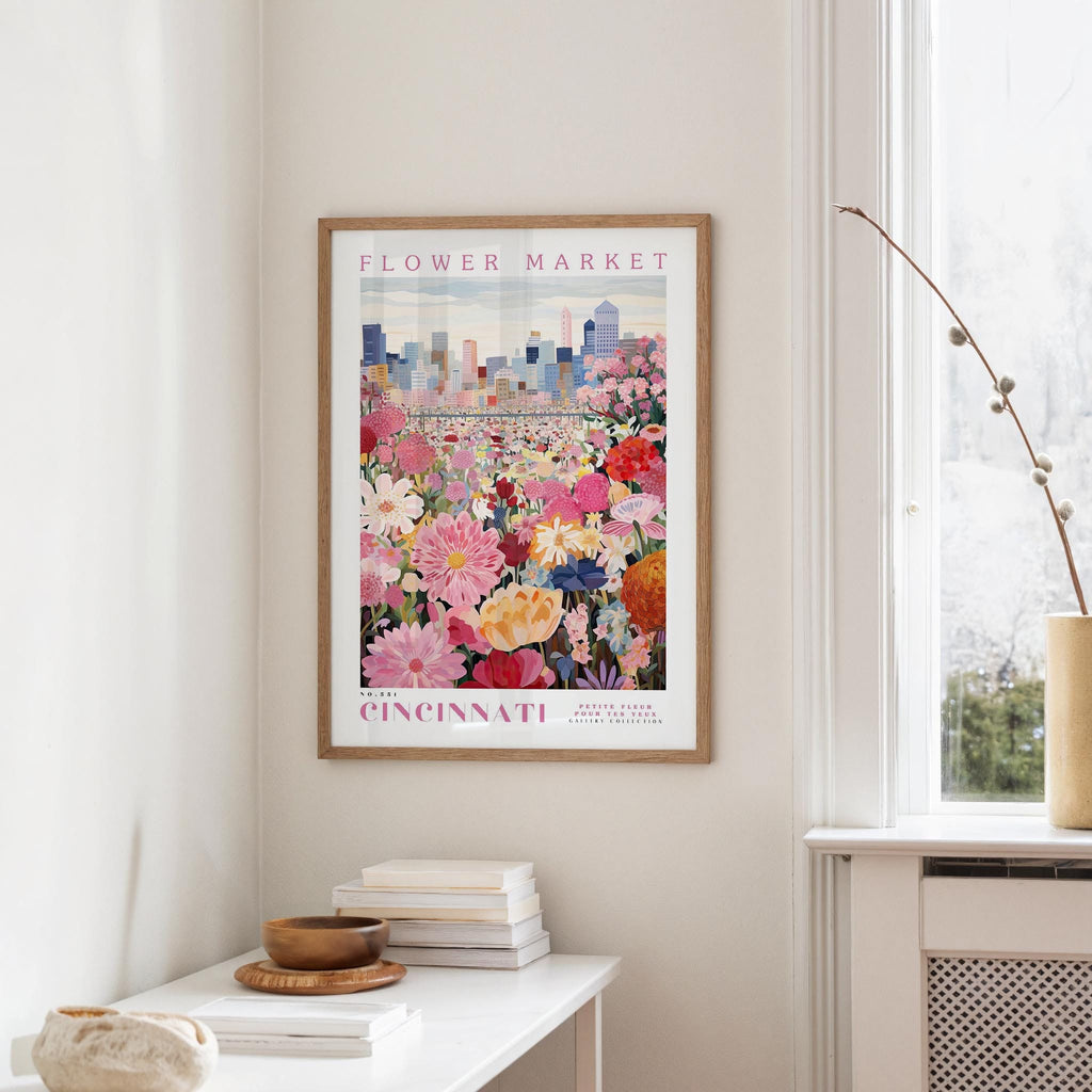 a framed poster of a colorful flower market in Cincinnati, Ohio, hanging on a wall in a room with a window and a small table with books and a vase.