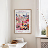 a framed poster of a colorful flower market in Cincinnati, Ohio, hanging on a wall in a room with a window and a small table with books and a vase.