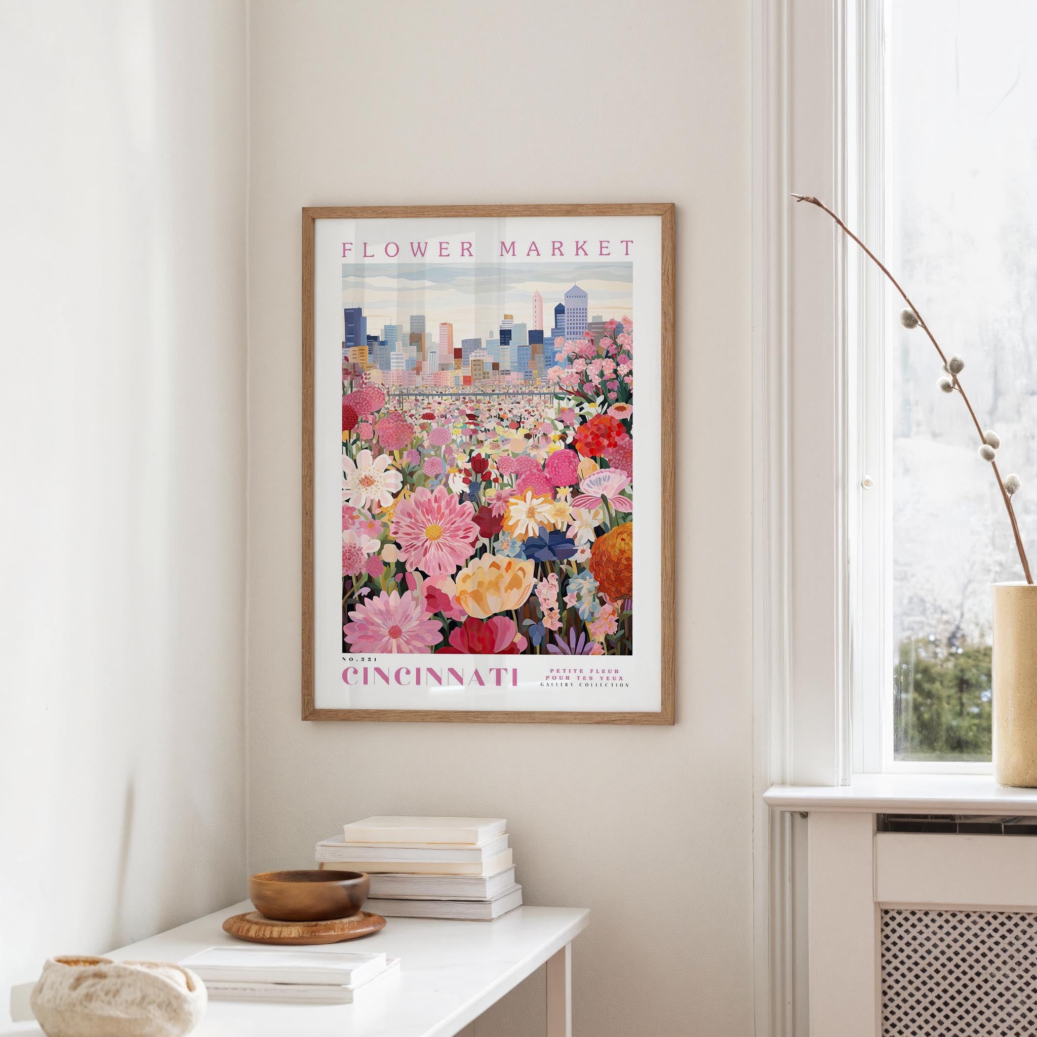 a framed poster of a colorful flower market in Cincinnati, Ohio, hanging on a wall in a room with a window and a small table with books and a vase.