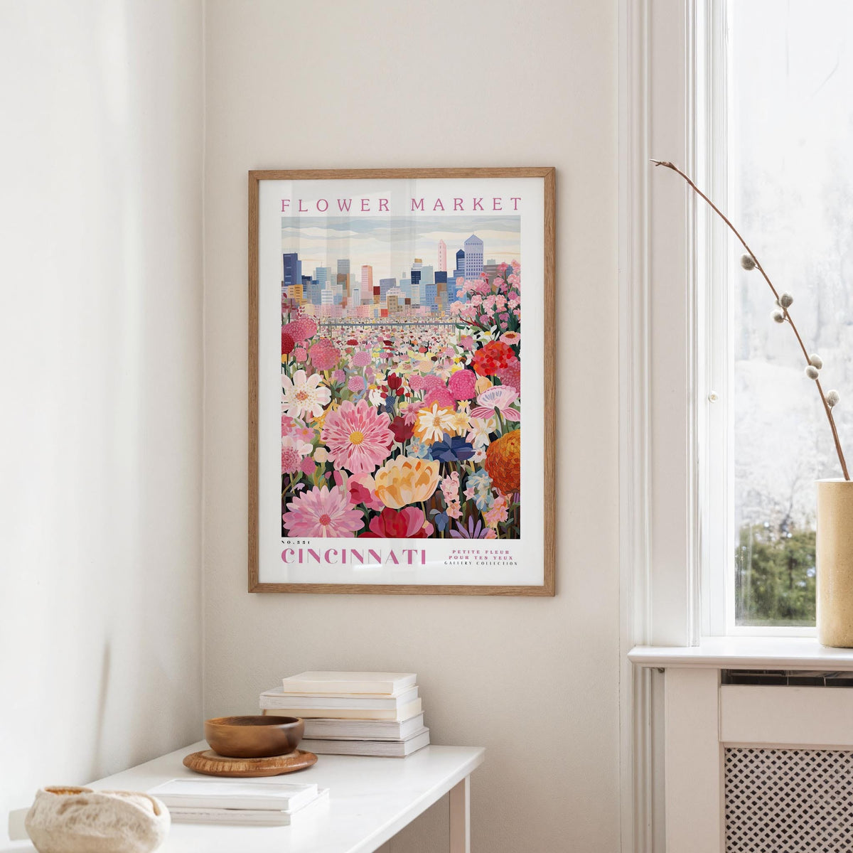 a framed poster of a colorful flower market in Cincinnati, Ohio, hanging on a wall in a room with a window and a small table with books and a vase.