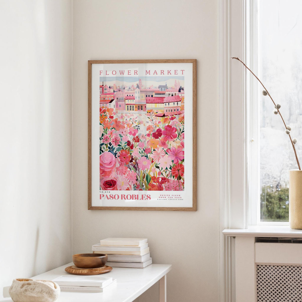 a framed poster of a colorful floral scene hanging on a wall, with a white table and a vase of flowers in the foreground.