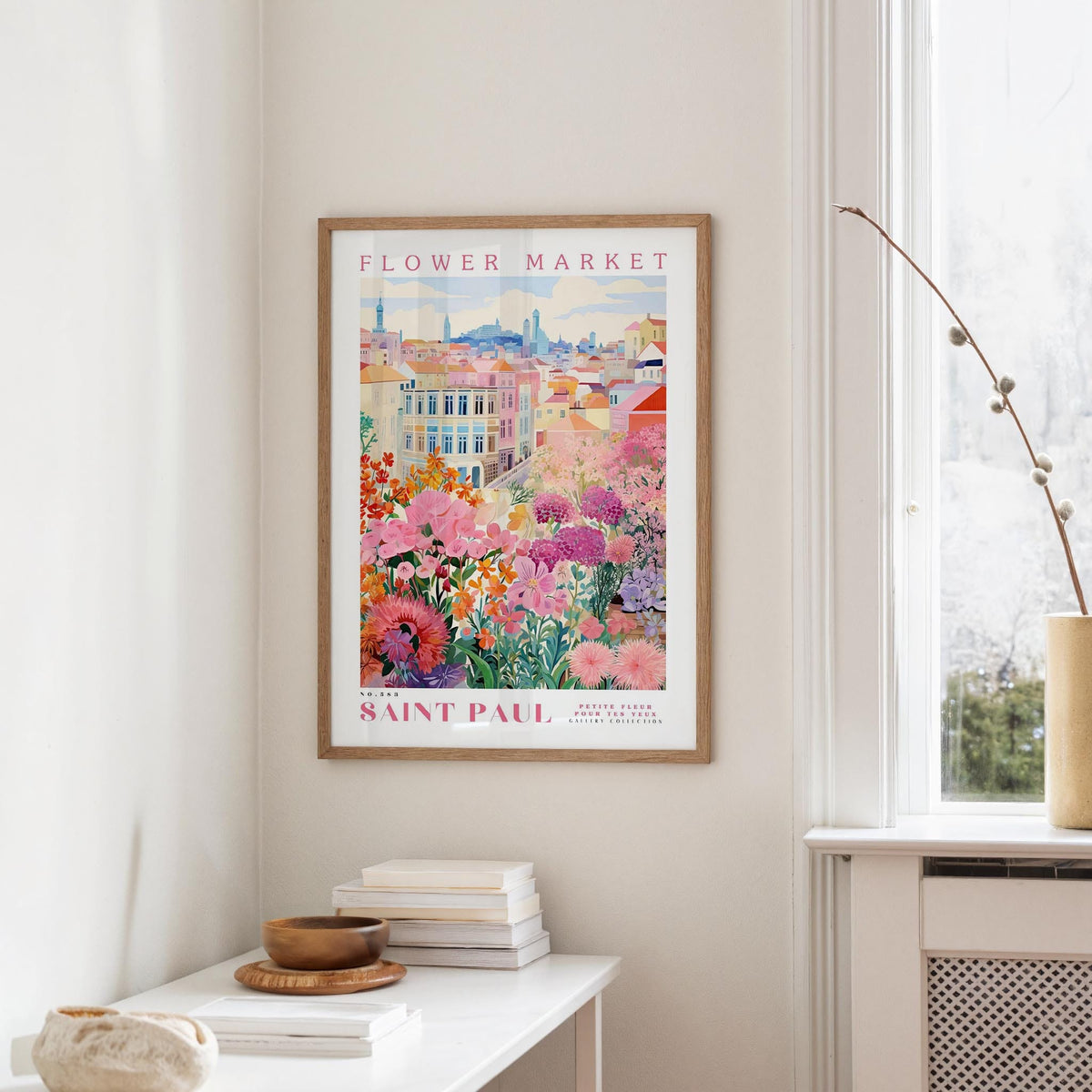 a framed poster of a colorful floral scene hanging on a wall, with a window and a small table with books and a vase in the foreground.