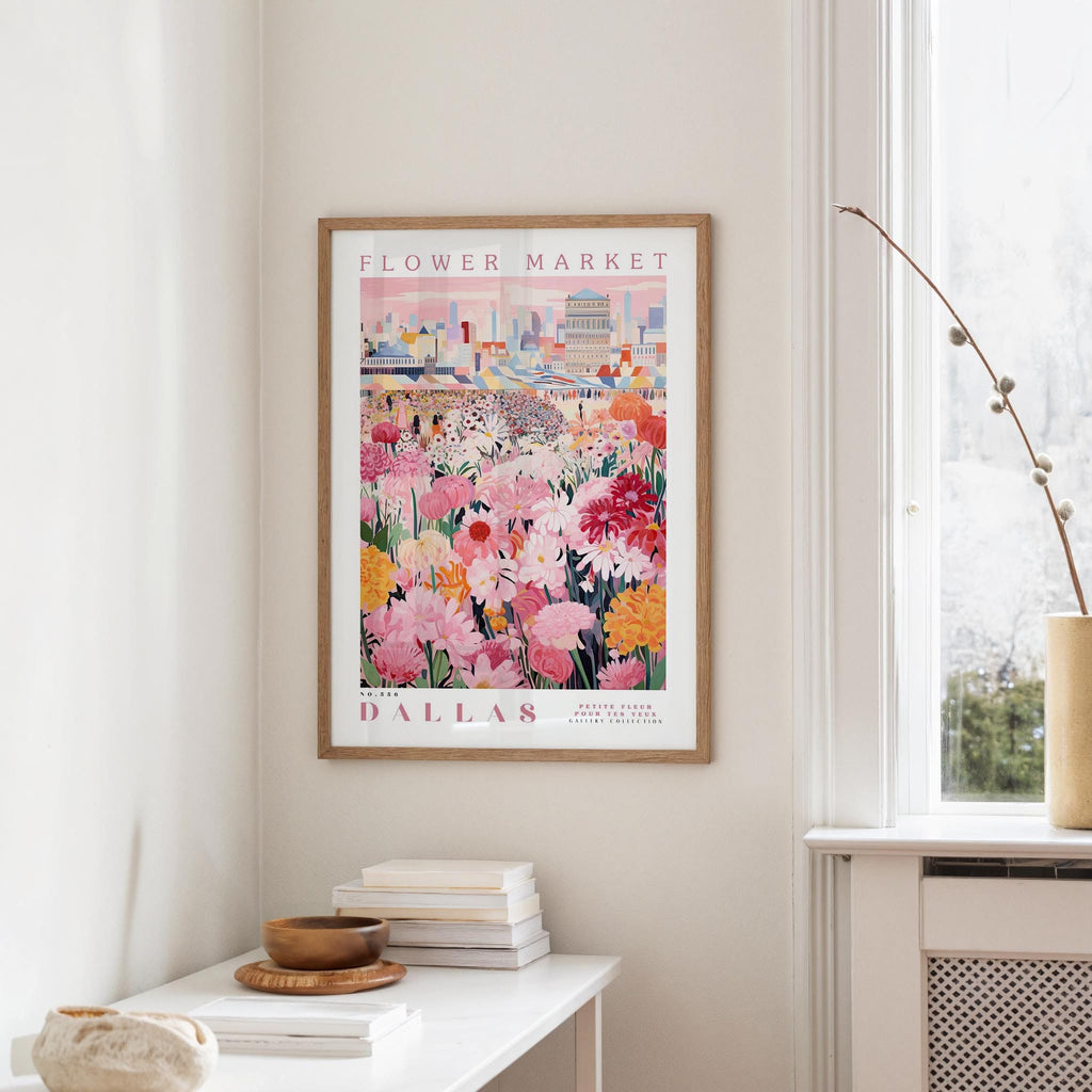a framed poster of a colorful floral scene hanging on a wall, with a white table and a vase of flowers in the foreground.