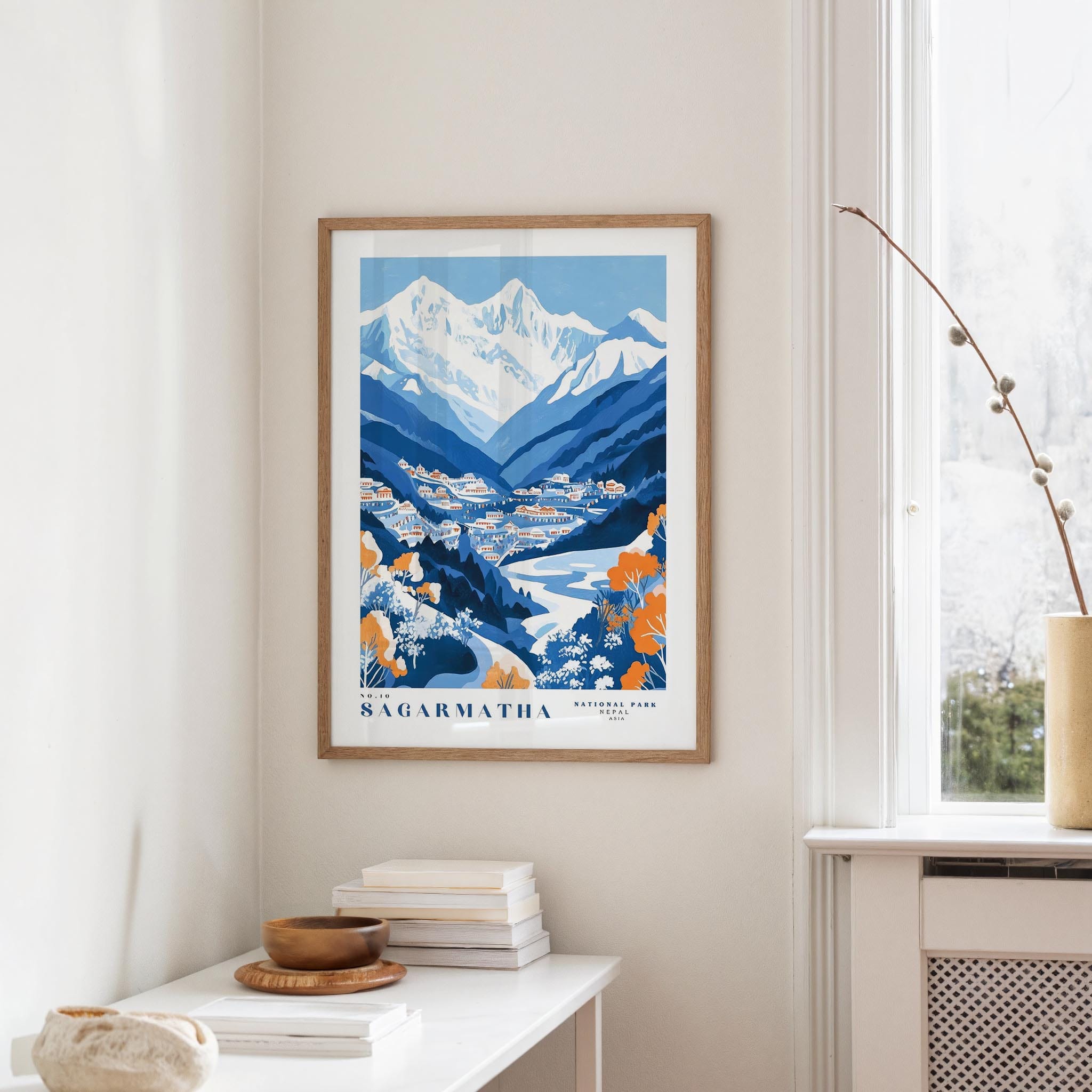 a framed poster of a mountainous landscape with a river, hanging on a wall in a room with a white table and a window.