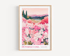 A framed poster depicting a scenic landscape with a field of pink flowers, mountains in the background, and a bridge in the distance.