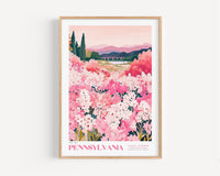 A framed poster depicting a scenic landscape with a field of pink flowers, mountains in the background, and a bridge in the distance.