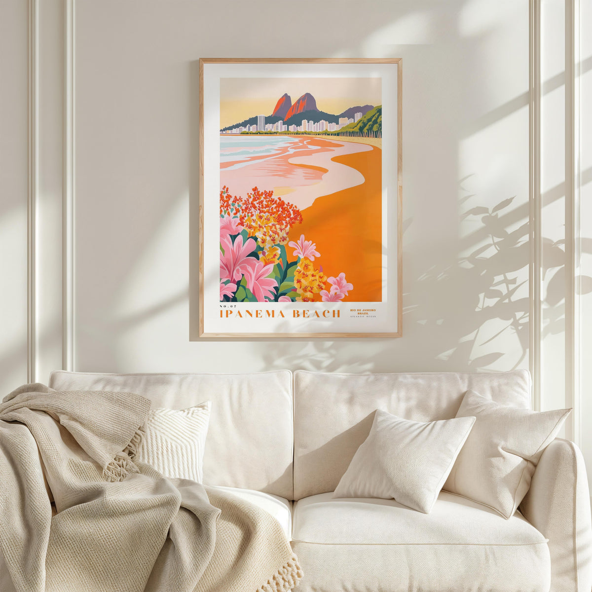 a framed poster of a beach scene with mountains in the background, hanging on a wall above a white couch with a blanket draped over it.
