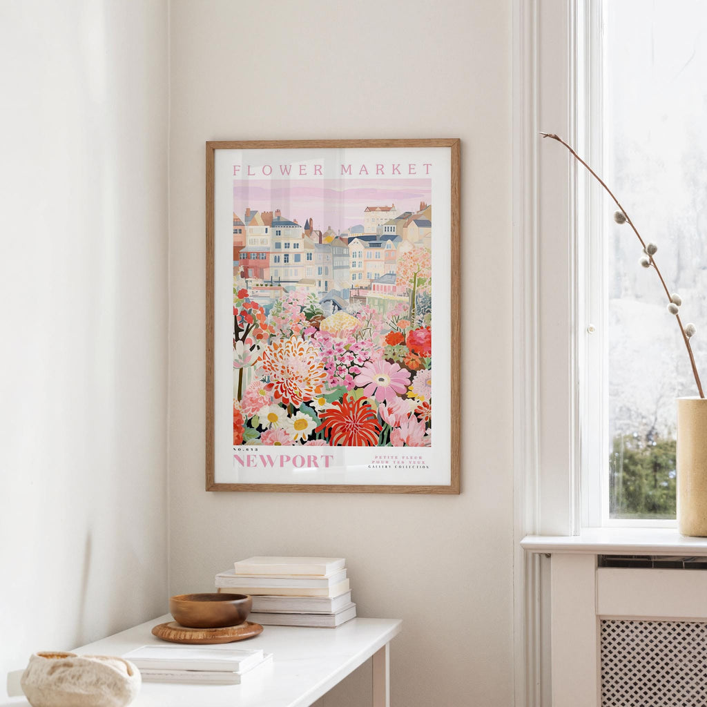 a framed poster of a colorful floral scene hanging on a wall, with a white table and a vase of flowers in the foreground.