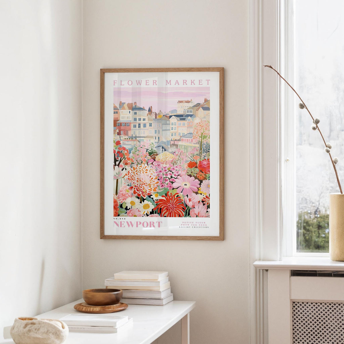 a framed poster of a colorful floral scene hanging on a wall, with a white table and a vase of flowers in the foreground.