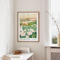 a framed poster of a scenic landscape with a field of flowers, including white and pink flowers, and a building in the background. The poster is hanging on a wall in a room with a window and a small table with books and a vase.