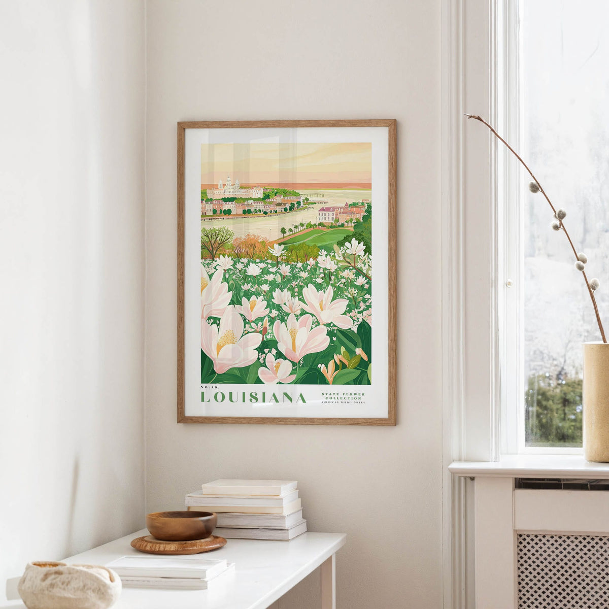 a framed poster of a scenic landscape with a field of flowers, including white and pink flowers, and a building in the background. The poster is hanging on a wall in a room with a window and a small table with books and a vase.