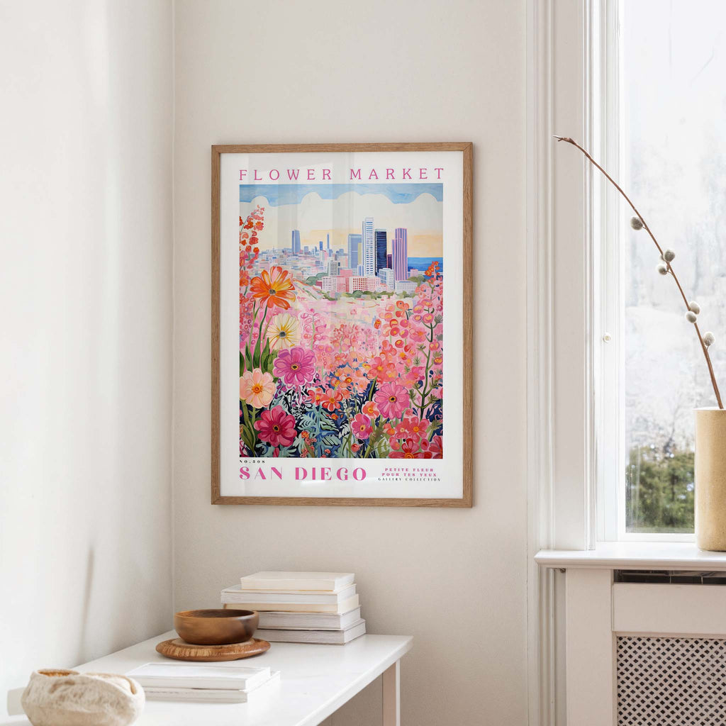 a framed poster of a colorful floral scene hanging on a wall, with a window and a small table with books and a vase in the foreground.