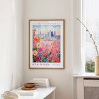 a framed poster of a colorful floral scene hanging on a wall, with a window and a small table with books and a vase in the foreground.