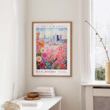 a framed poster of a colorful floral scene hanging on a wall, with a window and a small table with books and a vase in the foreground.