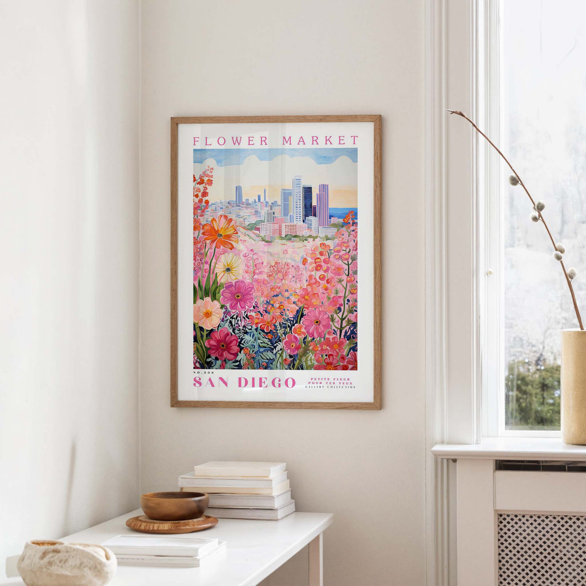a framed poster of a colorful floral scene hanging on a wall, with a window and a small table with books and a vase in the foreground.