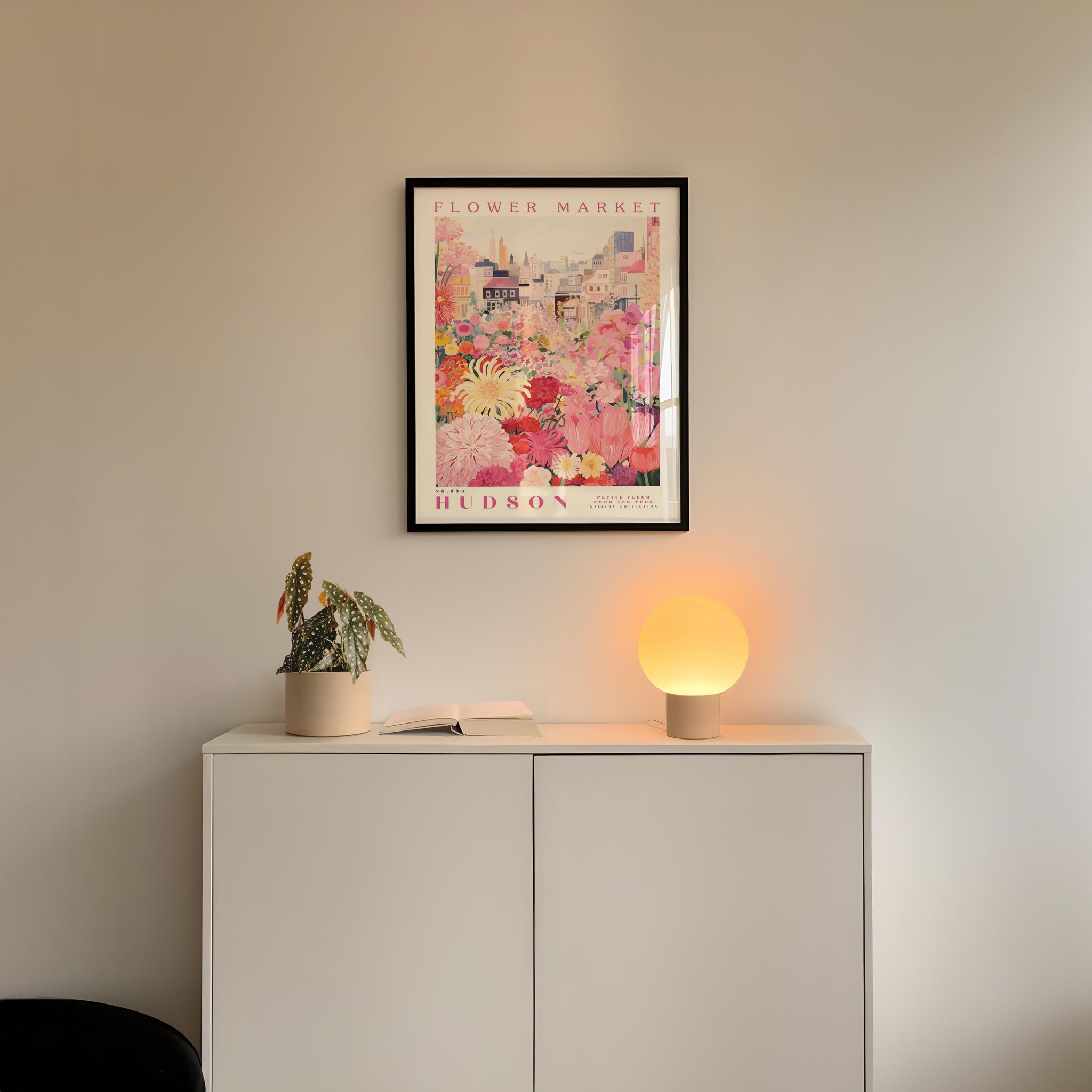 a white cabinet with a potted plant and a lamp on top, set against a wall with a framed poster of a flower market above it.