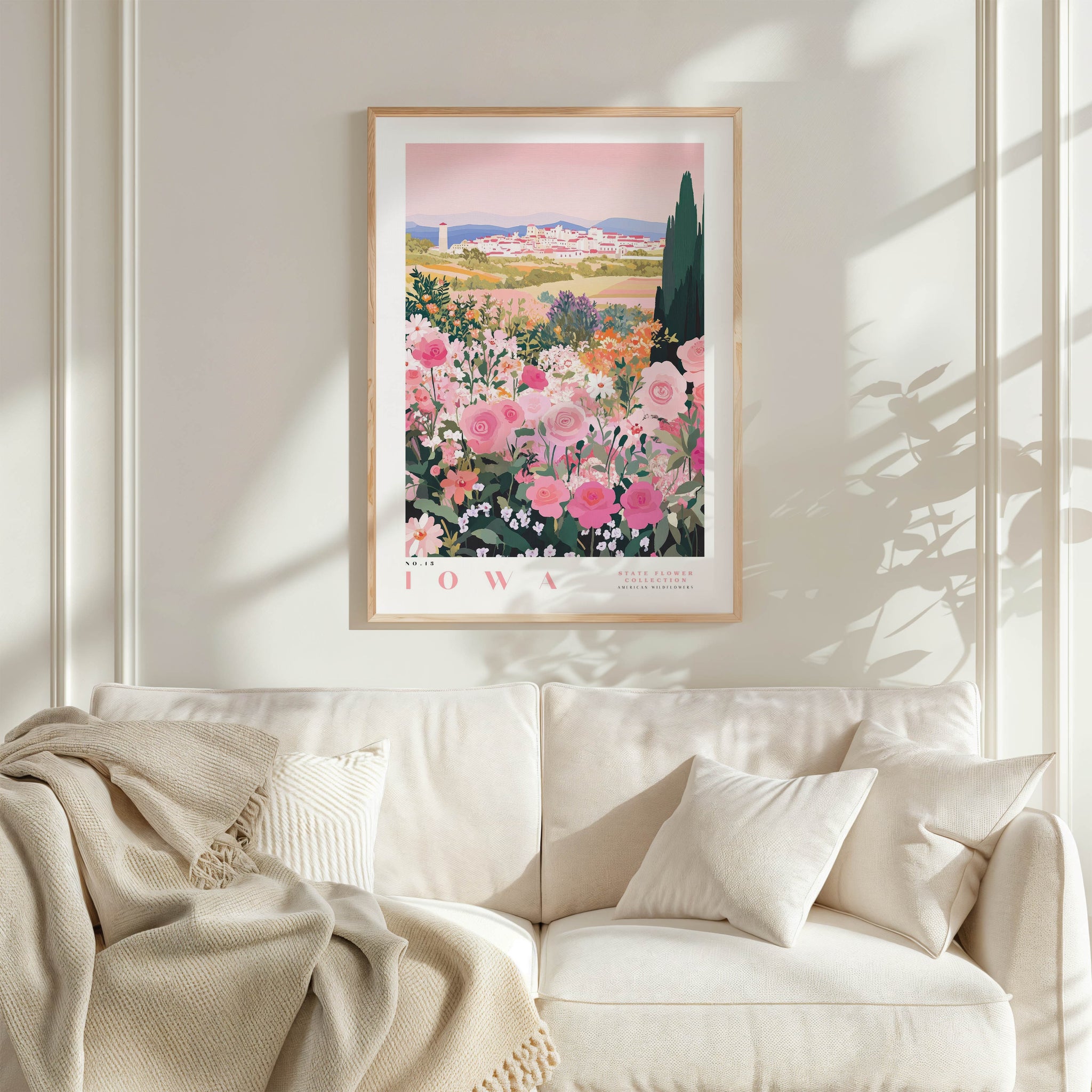 a framed poster of a garden with pink flowers hanging on a wall above a white couch with pillows.