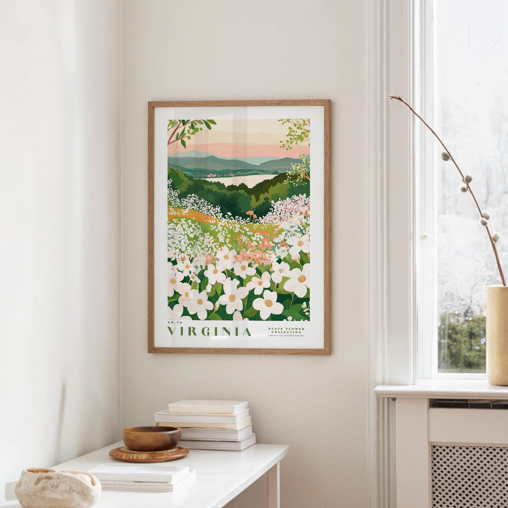 a framed poster of a scenic landscape with a field of flowers, hanging on a wall in a room with a white table and a window.