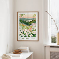 a framed poster of a scenic landscape with a field of flowers, hanging on a wall in a room with a white table and a window.