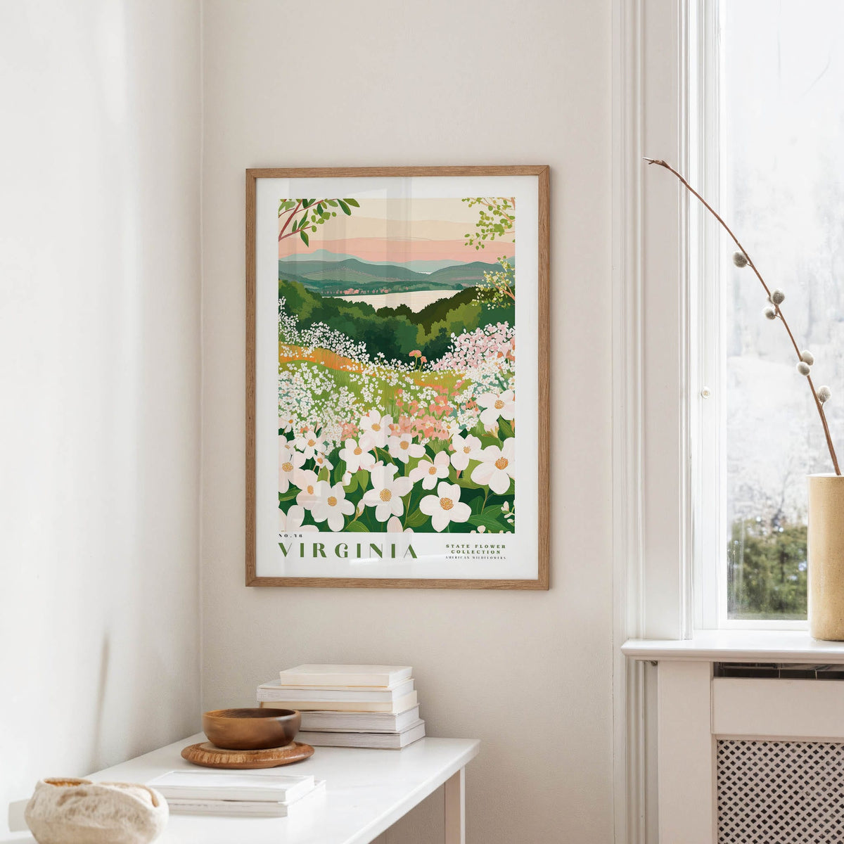 a framed poster of a scenic landscape with a field of flowers, hanging on a wall in a room with a white table and a window.