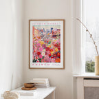 a framed poster of a colorful floral scene hanging on a wall, with a white table and a vase of flowers in the foreground.