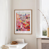 a framed poster of a colorful floral scene hanging on a wall, with a white table and a vase of flowers in the foreground.