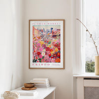 a framed poster of a colorful floral scene hanging on a wall, with a white table and a vase of flowers in the foreground.