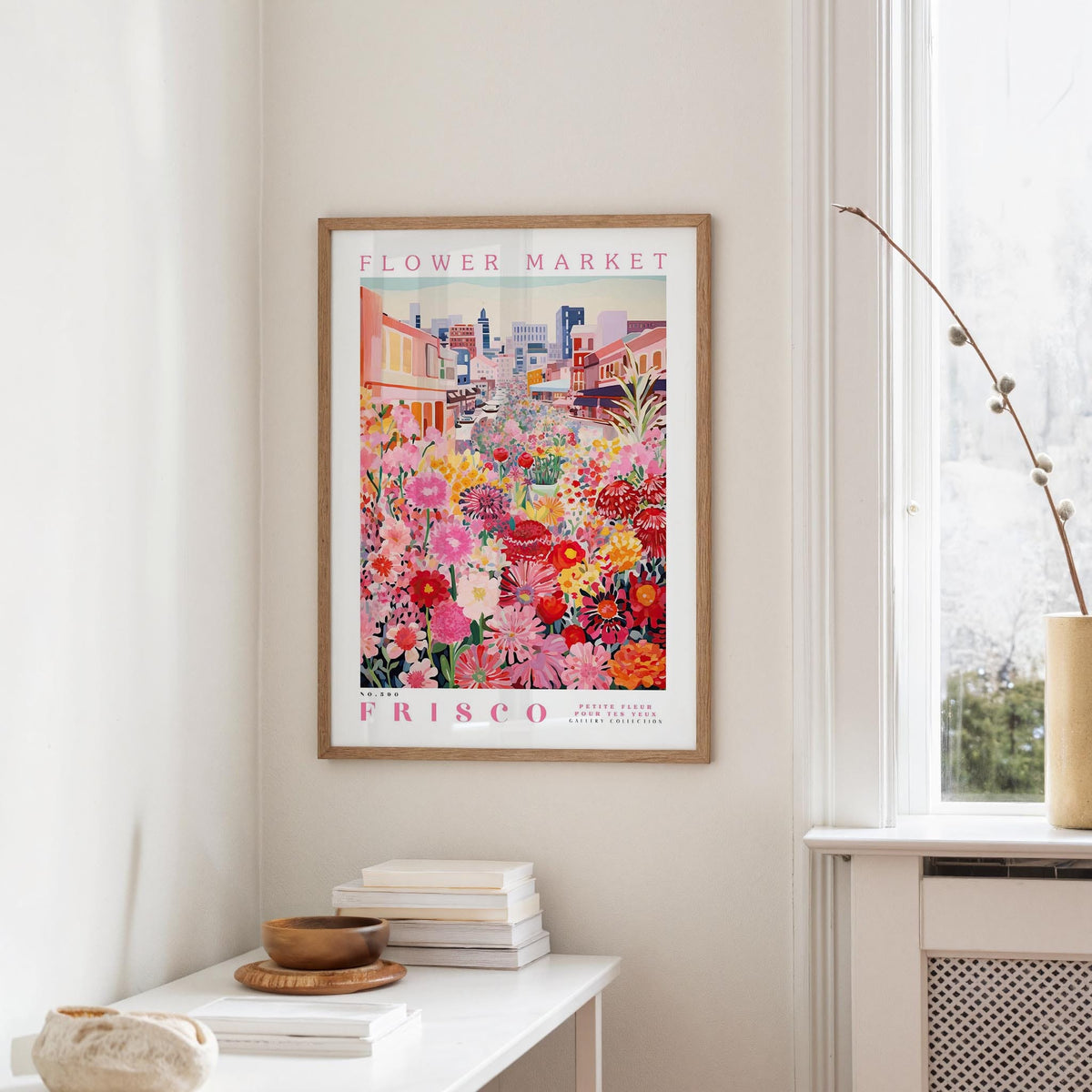 a framed poster of a colorful floral scene hanging on a wall, with a white table and a vase of flowers in the foreground.