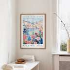 a framed poster of a colorful floral scene hanging on a wall, with a white table and a vase of flowers in the foreground.