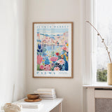 a framed poster of a colorful floral scene hanging on a wall, with a white table and a vase of flowers in the foreground.