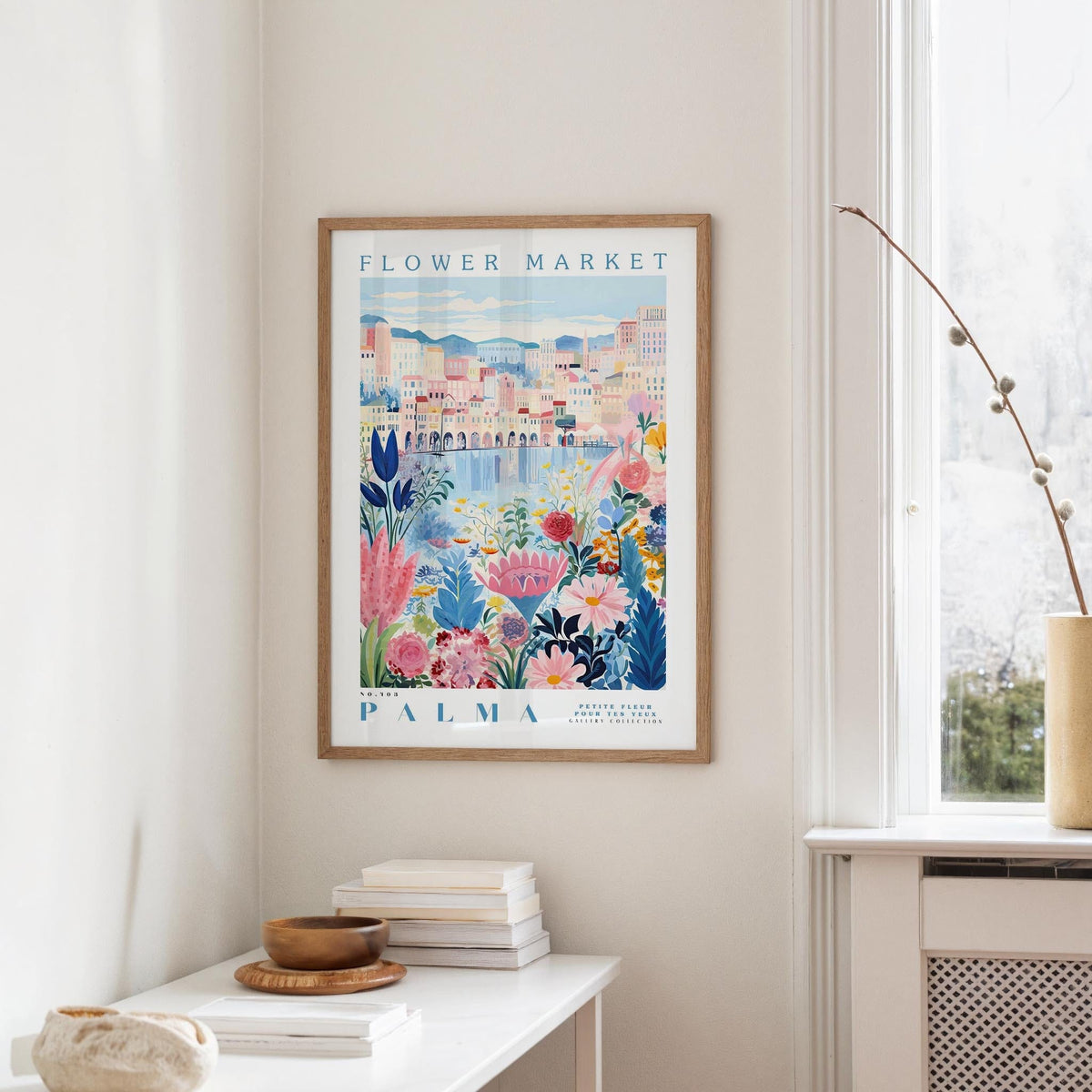 a framed poster of a colorful floral scene hanging on a wall, with a white table and a vase of flowers in the foreground.