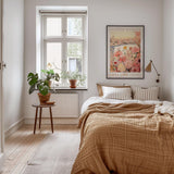 a cozy bedroom with a large bed, a wooden nightstand, and a framed poster of a flower market on the wall. The room has a warm, inviting atmosphere with a wooden floor and a rug.