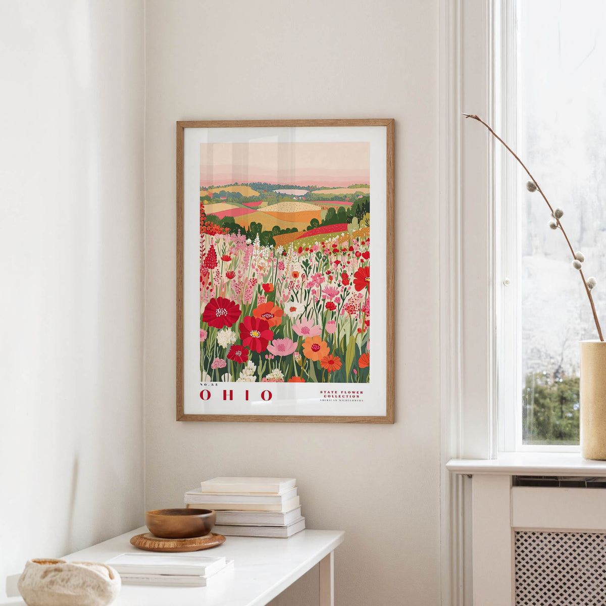 a framed poster of a colorful field of flowers hanging on a wall, with a white table and a vase of flowers in the foreground.